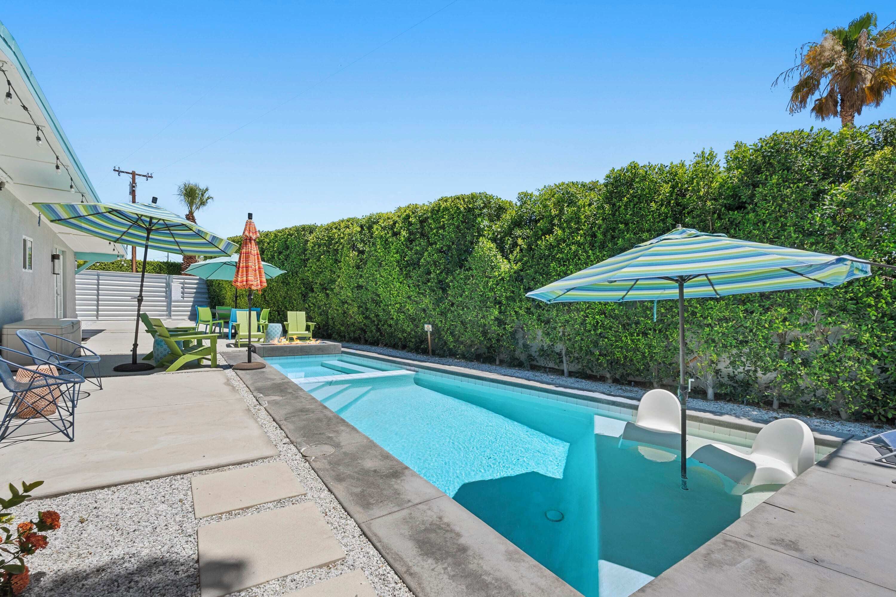 A swimming pool with lounge chairs and umbrellas, surrounded by a hedge and a house.
