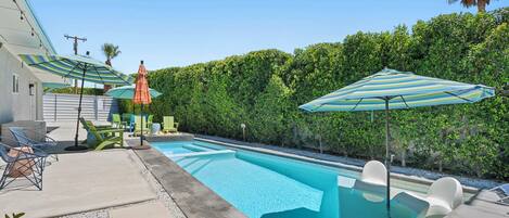 A swimming pool with lounge chairs and umbrellas, surrounded by a hedge and a house.