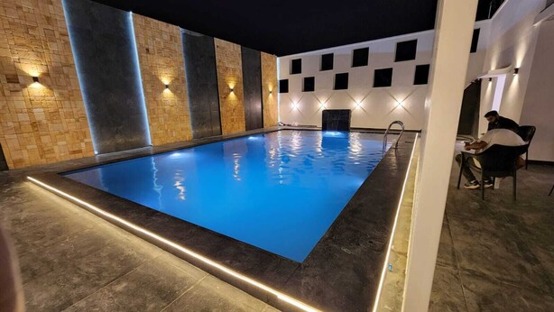 Ein rechteckiger, überdachter Pool mit dunkelblauem Wasser, umgeben von dunklem Boden und Wänden. Entlang des Randes befinden sich beleuchtete Wandleuchten, und im Hintergrund sitzt eine Person an einem Tisch.