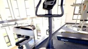 Una bicicleta estàtica a la gimnàstica, amb un pantalla digital i un banc de peses al fons.