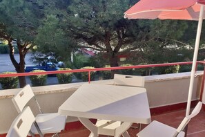 Ein Balkon mit einem weißen Tisch und Stühlen, einem roten Sonnenschirm und Blick auf Bäume und eine Straße.