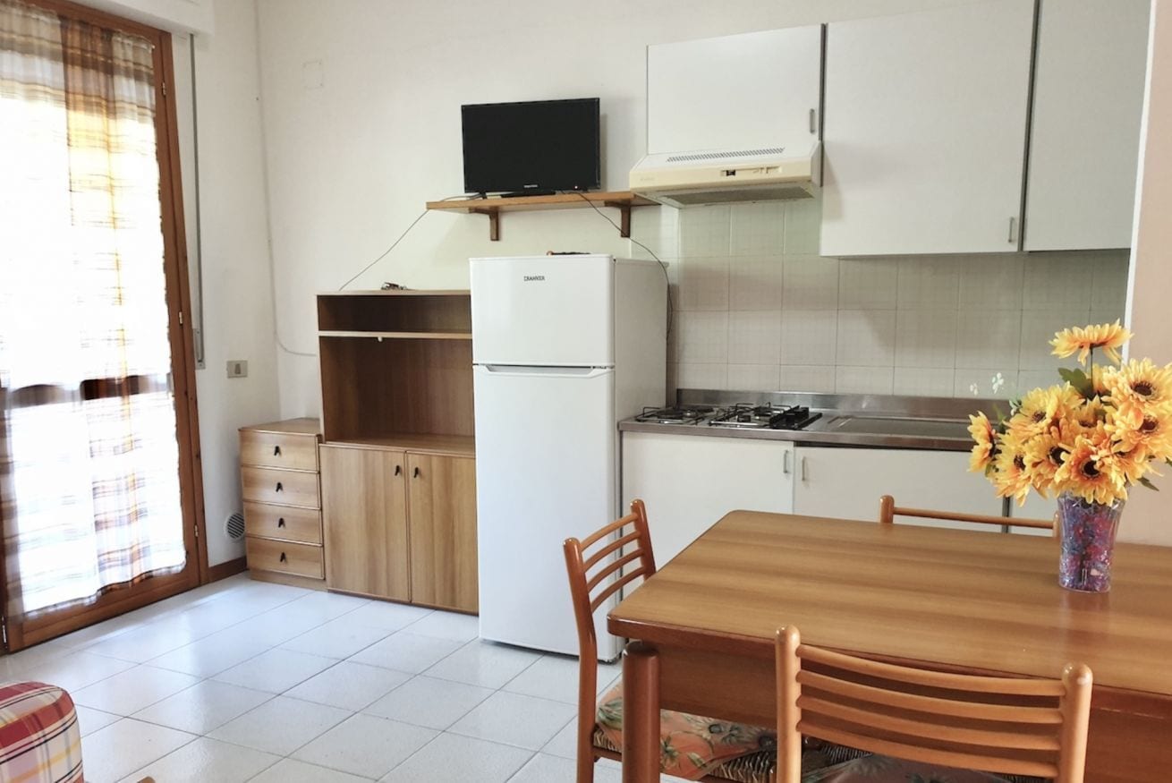 A compact kitchen with a refrigerator, stove, and wooden cabinets. There is a dining table with chairs and a vase with flowers.