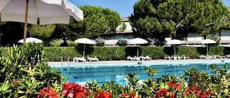 A pool area with white umbrellas and lounge chairs, surrounded by greenery and trees.