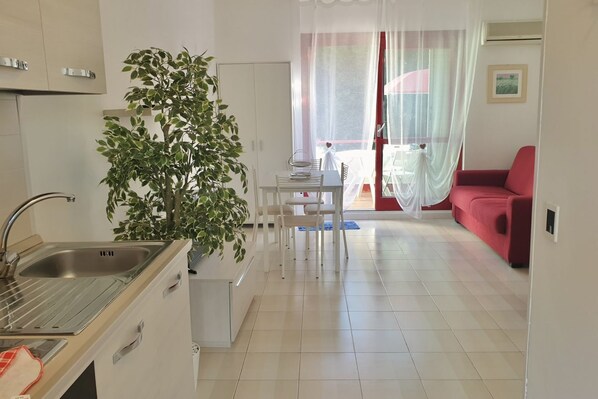 Cocina compacta con lavamanos, armarios y comedor con mesa y sillas. Una planta en maceta está en la esquina, y un sofá rojo se encuentra en la sala.