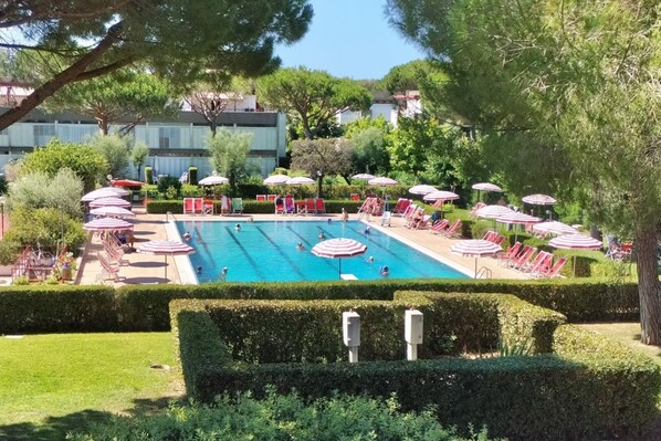 A swimming pool surrounded by red and white striped umbrellas and lounge chairs, with neatly trimmed hedges and trees in the background.