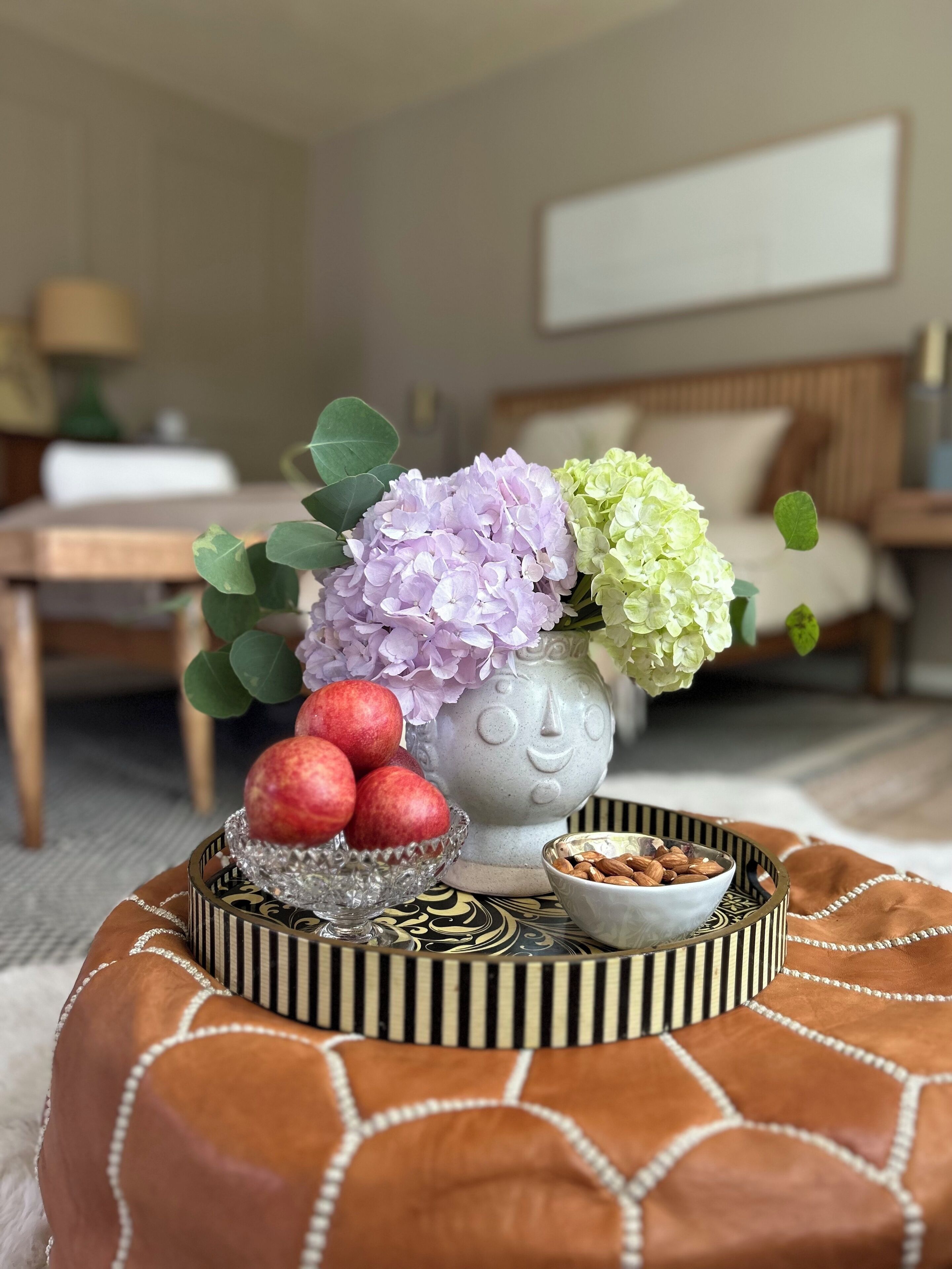 Una sala de estar acogedora con un pouf redondo, un jarrón con flores y un tazón de almendras.
