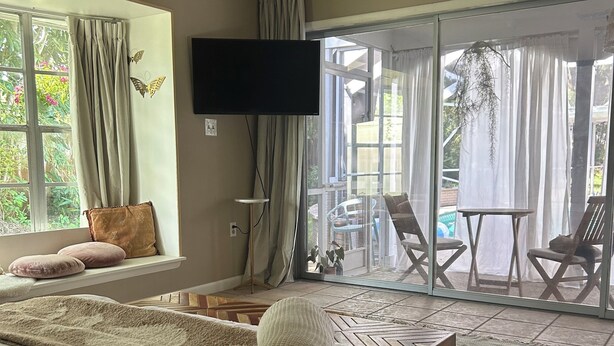 A bedroom with a large bed, a ceiling fan, a TV, and a sliding glass door leading to an outdoor area with chairs and a table.