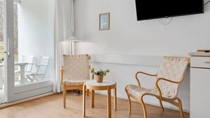 A modern room with a TV mounted on the wall, a wooden table, two woven chairs, a small potted plant, and a white door leading to an outdoor area.