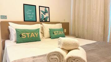 A hotel room with a bed, two pillows with 'STAY' text, a framed 'VIVE! Gai da do casulo STAY' sign, and a rolled towel on the bed.