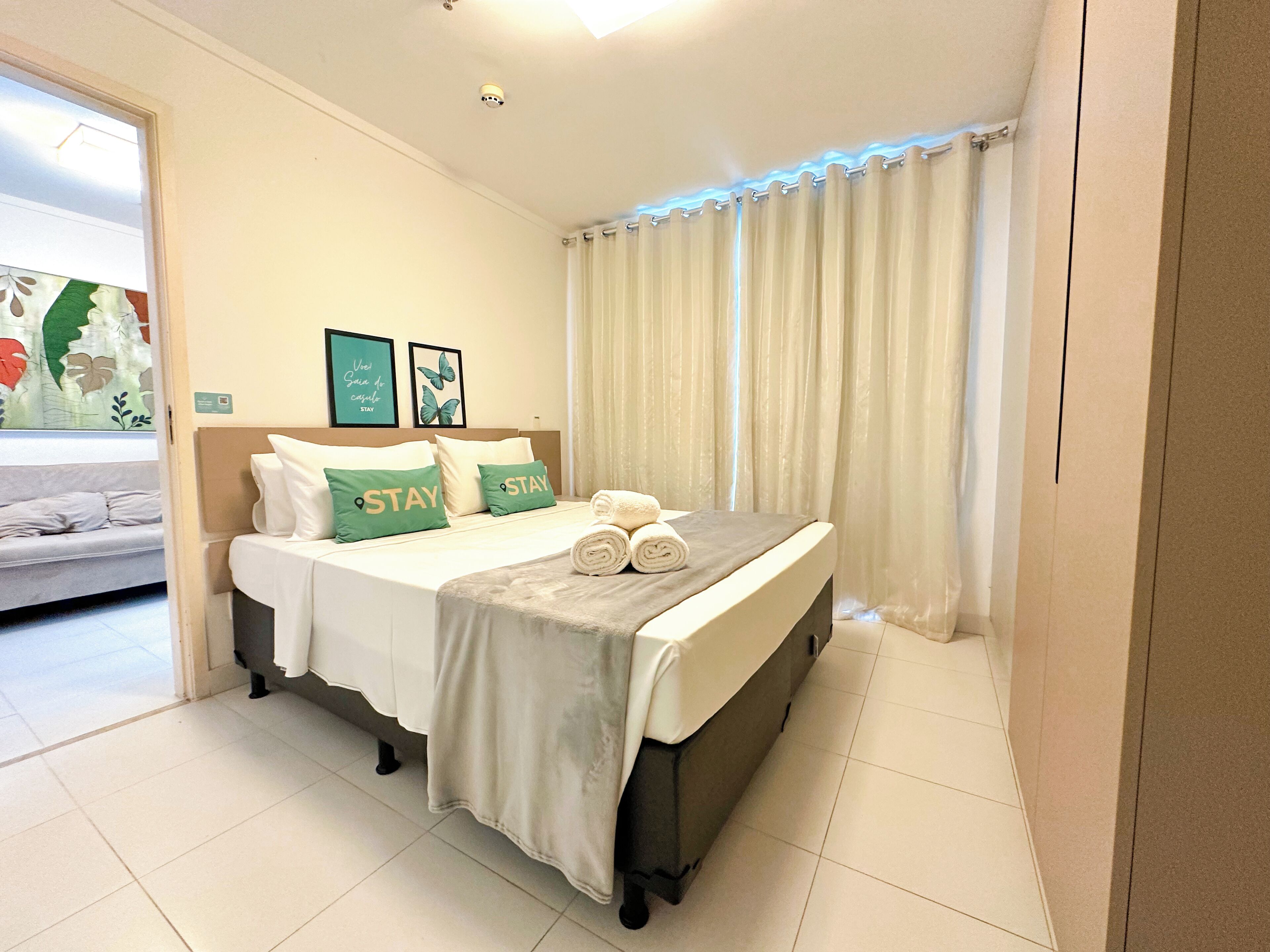 A hotel room with a bed, a sofa, a window with curtains, and a wall with framed pictures.