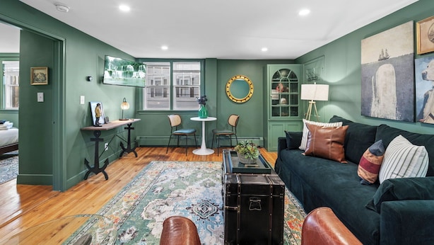 A living room with a green wall, a black trunk, a green sofa with multiple pillows, a patterned rug, and a television mounted on the wall.