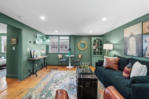Un salón con una pared verde, un baúl negro, un sofá verde con varios cojines, una alfombra estampada y un televisor montado en la pared.