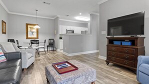 Un salon avec un canapé gris, une table basse sur laquelle se trouve un plateau, un téléviseur posé sur un meuble en bois, et une cuisine à l’arrière-plan.