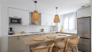 A modern kitchen with white cabinetry, a stainless steel refrigerator, a microwave, and a coffee maker. There are two hanging pendant lights and a wooden island with chairs.