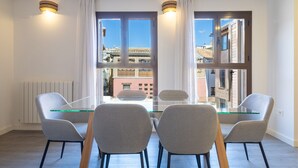 Un espace repas moderne avec une table en verre et quatre chaises, de grandes fenêtres avec des rideaux et une vue sur les bâtiments environnants.