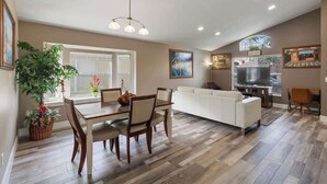 A modern living room with a white sofa, dining table, and chairs. There is a TV, a fireplace, and a large window.