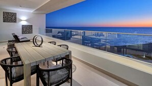 Un balcón moderno con vista al océano al atardecer, que incluye una mesa y sillas para comer, y decoración en la pared.