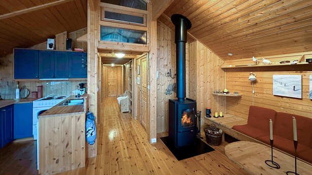 A cozy cabin interior with a central wood-burning stove, a kitchenette, and a seating area with a red sofa and a table. - 3 bedroom cozy home in Tjørhom (Tjørhom)