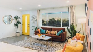 A living room with a brown sofa, yellow armchair, and a coffee table. There is a large window with curtains, a potted plant, and a mirror on the wall. - Casa De Colores w/ Arcade, hot tub + more - J Tree

 (Joshua Tree)