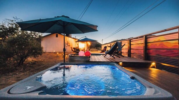 A hot tub with a deck, surrounded by outdoor furniture and umbrellas, with a building in the background. - Casa De Colores w/ Arcade, hot tub + more - J Tree

 (Joshua Tree)