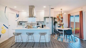 A modern kitchen with white cabinets, a central island with bar stools, and a dining area with a round table and chairs. - Casa De Colores w/ Arcade, hot tub + more - J Tree

 (Joshua Tree)