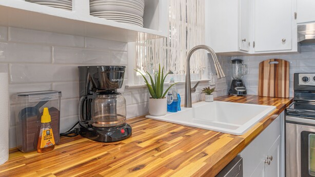 Una cocina con armarios blancos, encimera de madera, cafetera, fregadero y estufa.