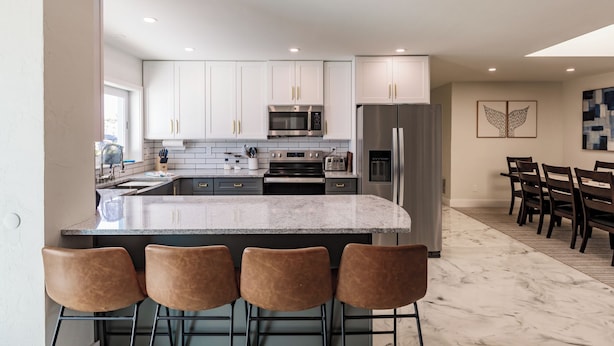 A modern kitchen with white cabinets, a large island with bar stools, stainless steel appliances, and a dining area with a table and chairs. - Family Escape w/ 5 bedrooms, pool, hot tub & canal (Cape Coral)
