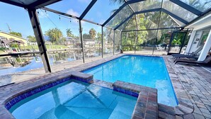 A swimming pool with a glass enclosure, surrounded by a patio area with a view of palm trees and a building in the background. - Family Escape w/ 5 bedrooms, pool, hot tub & canal (Cape Coral)