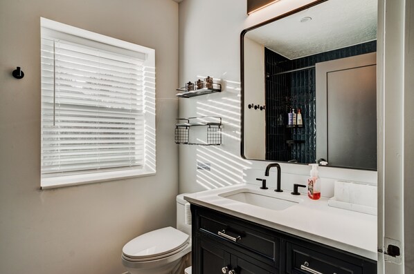 A bathroom with a toilet, a sink, a mirror, and a window with blinds.
