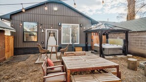 A rustic cabin with a large wooden table, two chairs, a hot tub, and string lights.