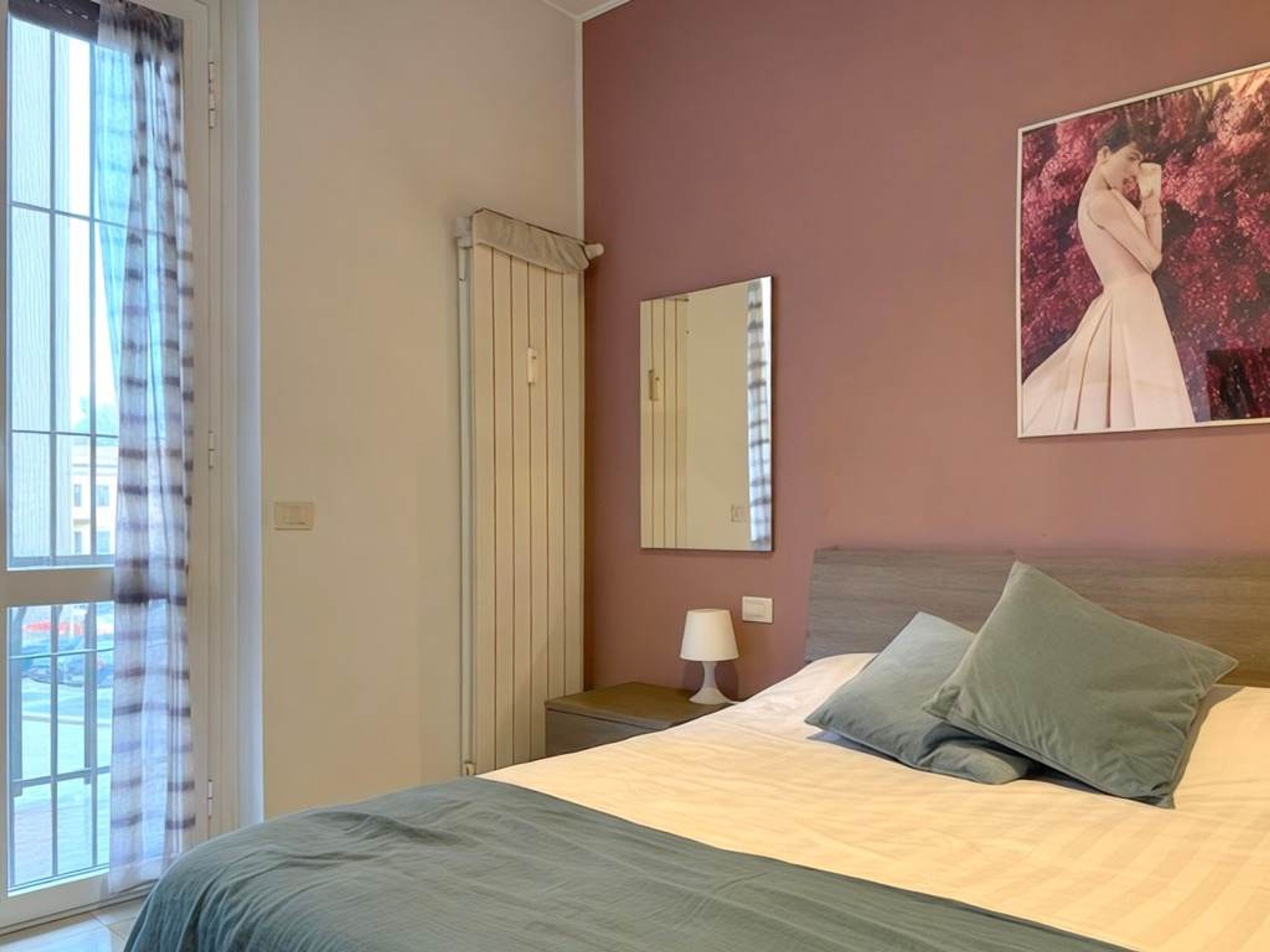 A bedroom with a bed, a nightstand, a lamp, a window with curtains, a radiator, and a painting on the wall.