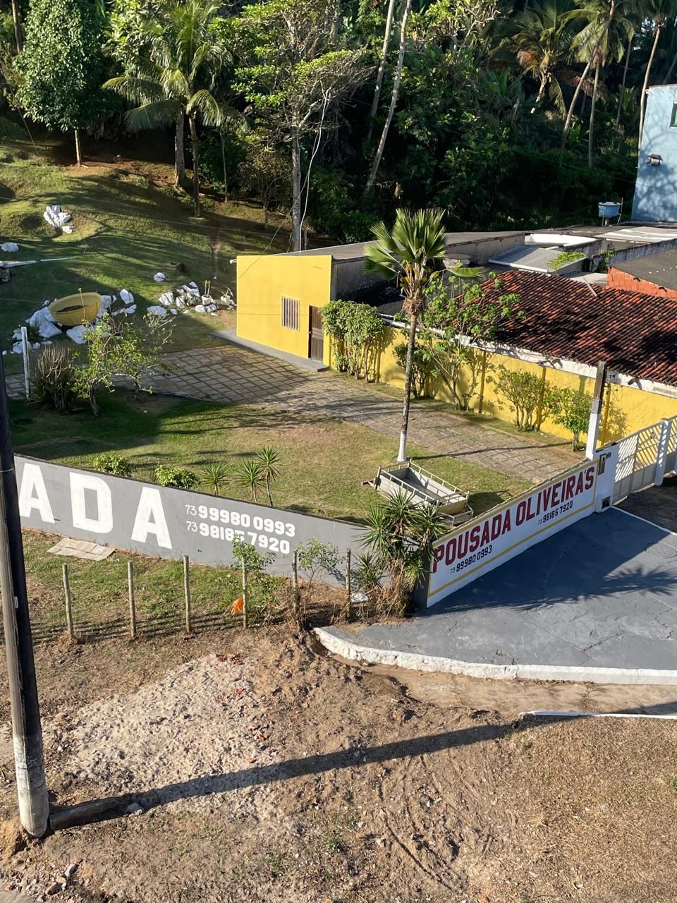 A residential area with a yellow building, a paved driveway, and a signboard with contact numbers.