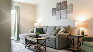 Una sala de estar moderna con un sofá gris, dos mesitas de noche con lámparas, una mesa de centro con libros y un sillón verde.