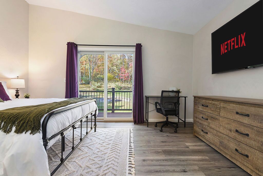 A bedroom with a bed, a desk, a dresser, a TV, and a balcony with curtains.