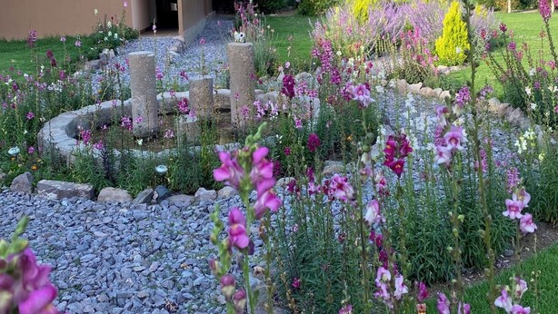Een rustiek gebouw met pannendak, omgeven door een verzorgde tuin met verschillende bloeiende planten en een grindpad.