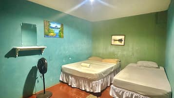 A room with two beds, a standing fan, a shelf with a mirror, and a painting on the wall.