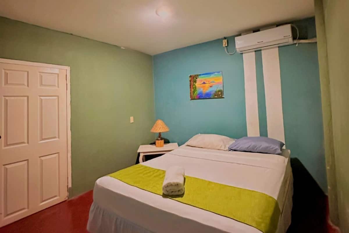 A hotel room with a bed, a nightstand, an air conditioning unit, and a door.