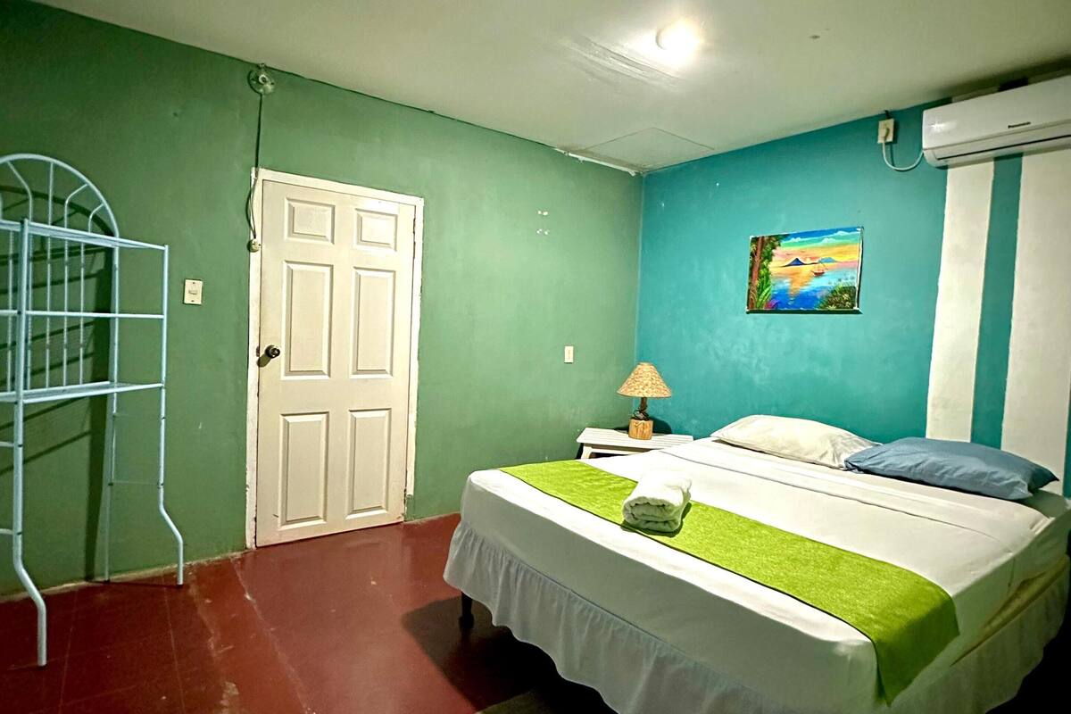 A bedroom with a bed, a nightstand, a lamp, a painting on the wall, and a white door.