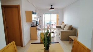 Una sala moderna con un sofá, una mesa de comedor con sillas, una zona de cocina con armarios y un televisor, además de un ventanal que proporciona luz natural.