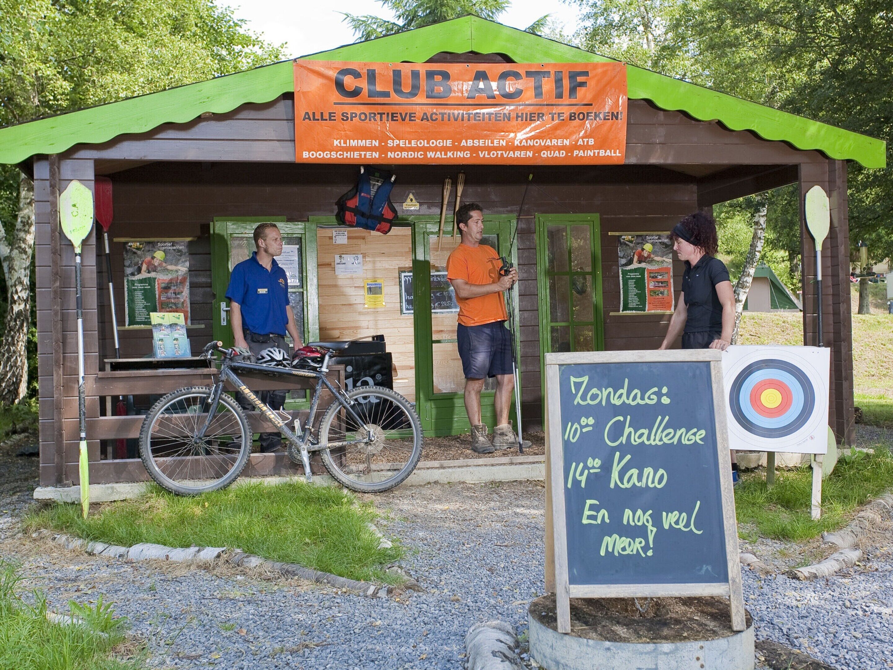 Eine kleine Holzhütte mit einem Schild mit der Aufschrift 'CLUB ACTIV' und einer Kreidetafel, die eine 10. Herausforderung und ein 14. Kanu-Event bewirbt.