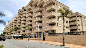 Un edificio comercial de varios pisos con numerosos balcones, un concesionario de vehículos comerciales y palmeras a lo largo de la acera.