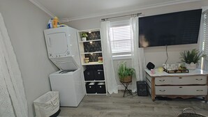 A laundry room with a stacked washer and dryer, shelves with storage bins, a potted plant, and a television on the wall. - Charming Cozy Cottage Pittsboro  (Pittsboro)