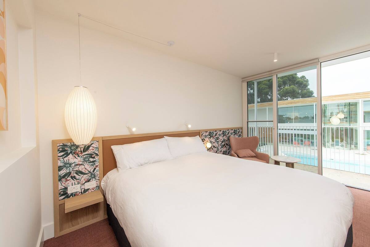 A modern hotel room with a large bed, a nightstand with a lamp, a chair, and a balcony with a pool view.