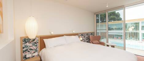 A modern hotel room with a large bed, a nightstand with a lamp, a chair, and a balcony with a pool view.