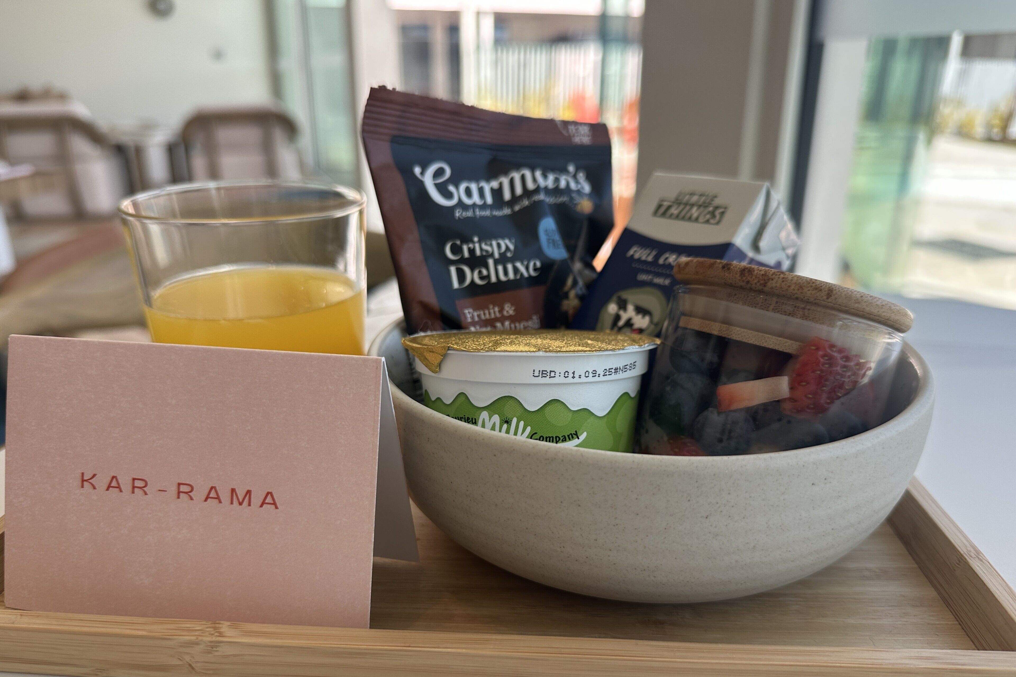 A breakfast tray with a glass of orange juice, a packet of Carmun's Crispy Deluxe, a container of milk, and a bowl with strawberries and yogurt.