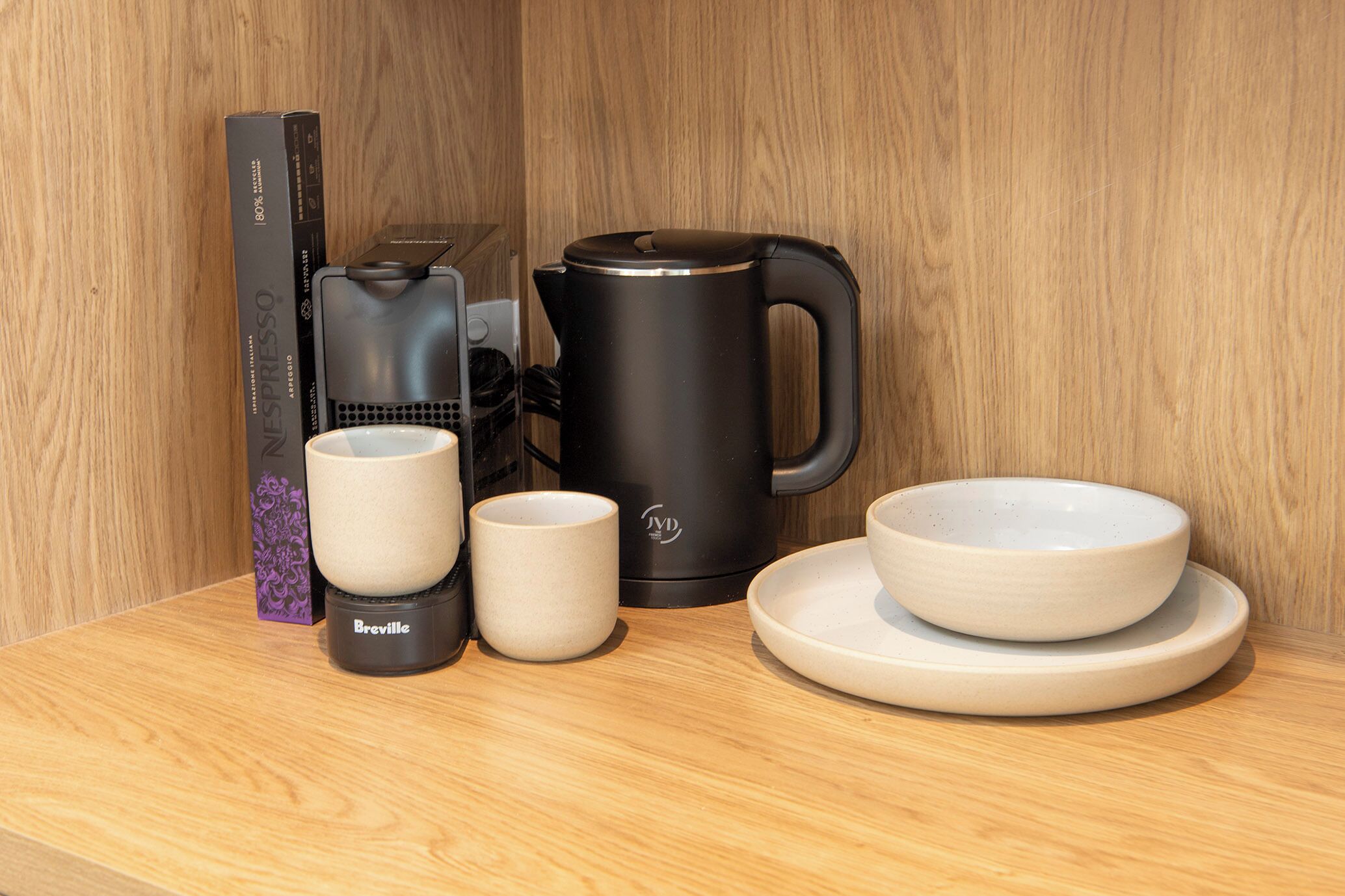 A coffee maker, two cups, a saucer, and a box on a wooden surface.