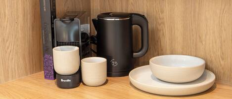 A coffee maker, two cups, a saucer, and a box on a wooden surface.