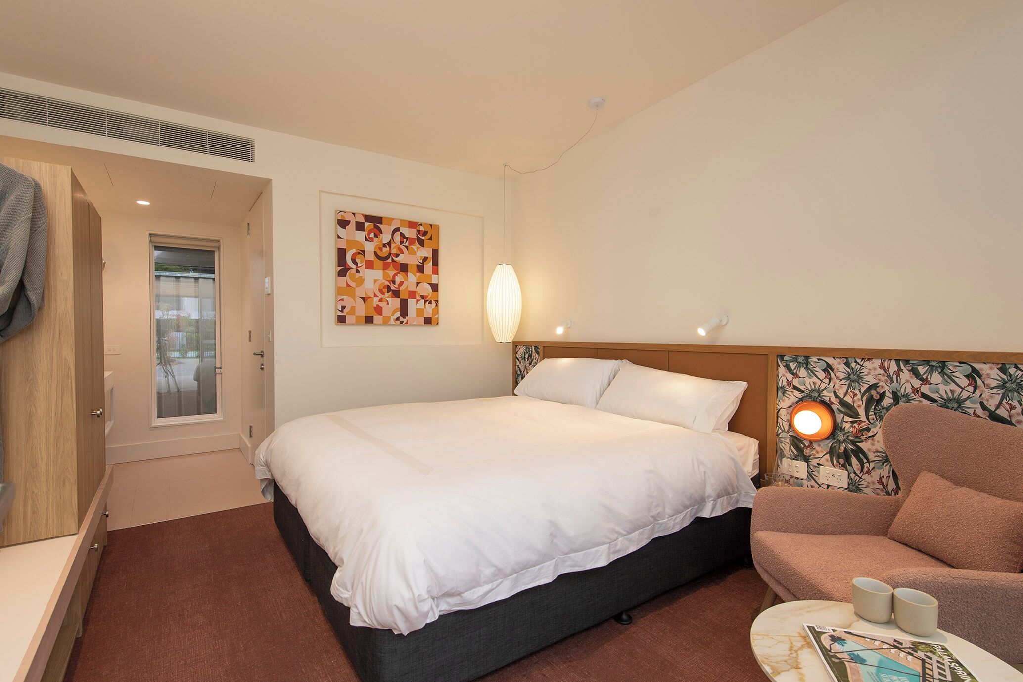 A hotel room with a bed, a chair, a table with two cups, a painting on the wall, and a door leading to another room.