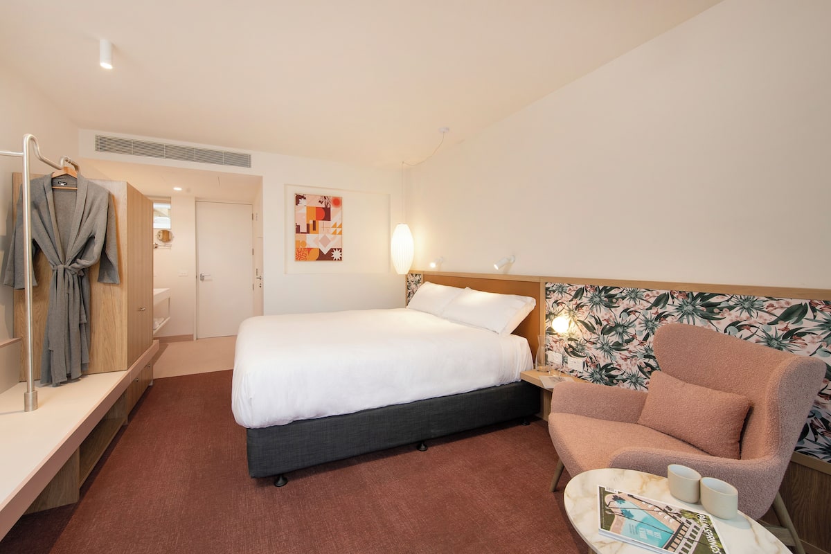 A hotel room with a bed, a chair, a small table with a magazine, and a robe hanging on a rack.