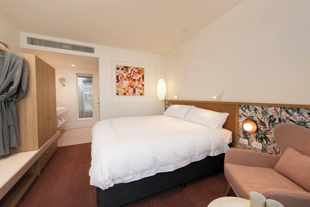 A hotel room with a bed, a sofa, a robe rack, a wall-mounted light, and a bathroom door.